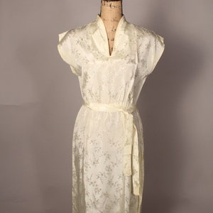 May include: A white vintage dress with a floral pattern and a sash tie at the waist. The dress has a V-neckline and short sleeves.