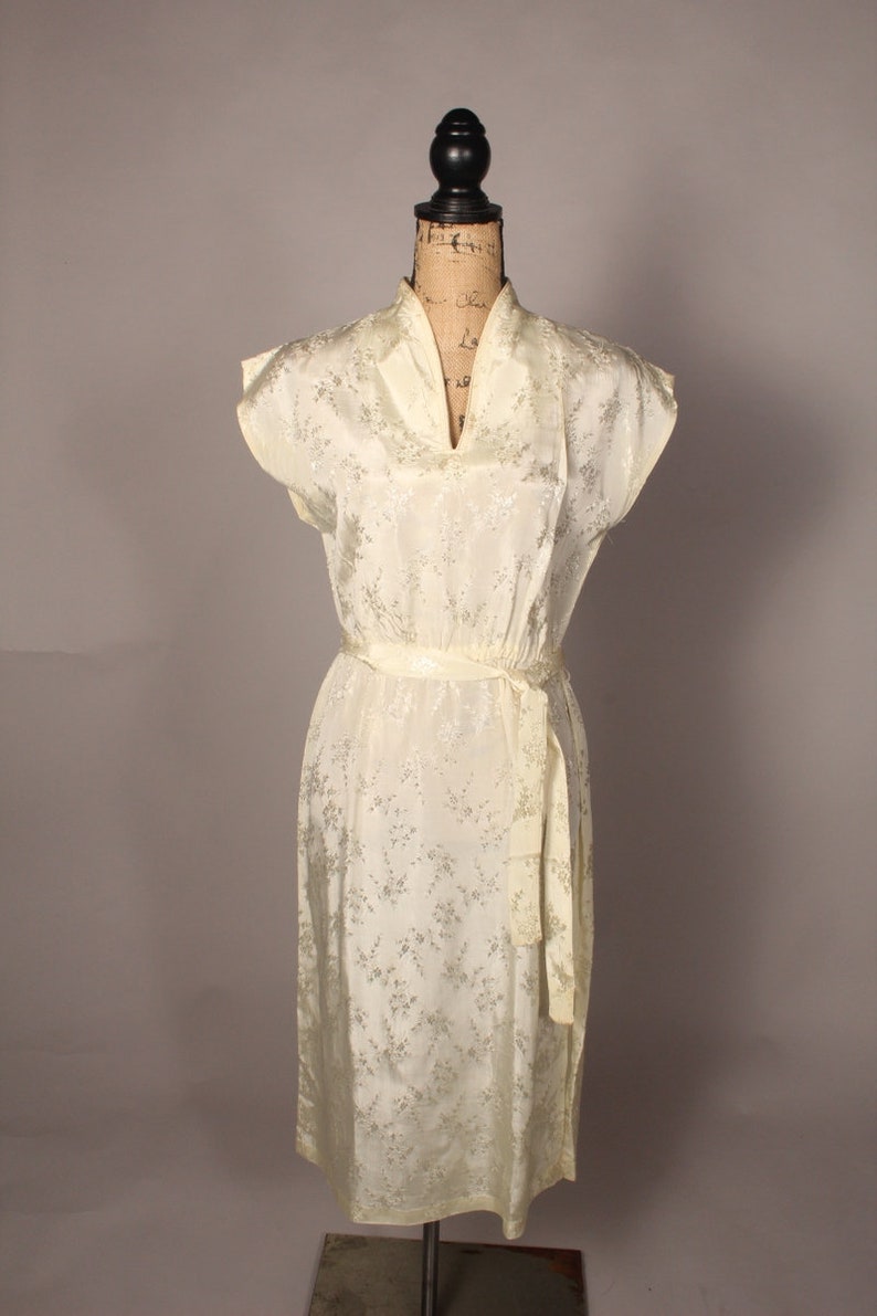 May include: A white vintage dress with a floral pattern and a sash tie at the waist. The dress has a V-neckline and short sleeves.