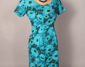 May include: A turquoise blue dress with a floral print. The dress has short sleeves and a fitted silhouette. It is on a mannequin wearing a dark blue beaded necklace.