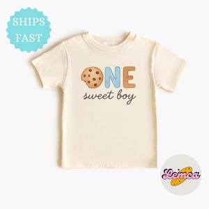 May include: Cream-colored toddler t-shirt with a cookie graphic and the words "ONE sweet boy". The word "ONE" is in blue and brown, and the cookie has a bite taken out of it.