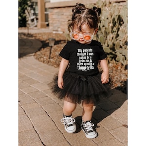 May include: A black short-sleeved t-shirt with white text that reads, My parents thought I was gonna be a princess & ended up with a thugerella, paired with a black layered tulle tutu skirt and black and white canvas sneakers.