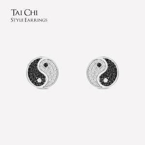 Può includere: Un paio di orecchini in stile Tai Chi. Ogni orecchino presenta un design yin e yang, con una metà riempita di pietre nere e l'altra di pietre bianche. Gli orecchini hanno un bordo argentato. Il testo "TAI CHI STYLE EARRINGS" è in alto.