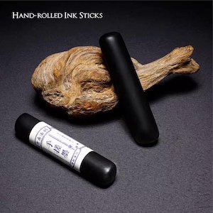 Puede incluir: Dos barras de tinta negras enrolladas a mano, una con una etiqueta blanca con caracteres asi&aacute;ticos, descansan sobre una superficie oscura con madera flotante. El texto "Hand-Rolled Ink Sticks" est&aacute; en la parte superior.