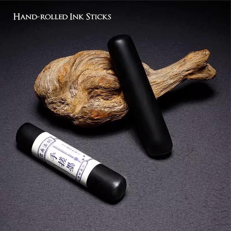 Puede incluir: Dos barras de tinta negras enrolladas a mano, una con una etiqueta blanca con caracteres asi&aacute;ticos, descansan sobre una superficie oscura con madera flotante. El texto "Hand-Rolled Ink Sticks" est&aacute; en la parte superior.