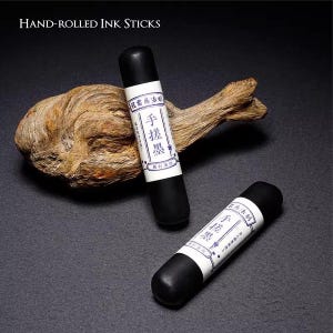 Puede incluir: Dos barras de tinta negras enrolladas a mano con etiquetas blancas con caracteres chinos. Las barras cil&iacute;ndricas descansan sobre una superficie oscura con un trozo de madera flotante. El texto "Hand-Rolled Ink Sticks" est&aacute; en la parte superior.