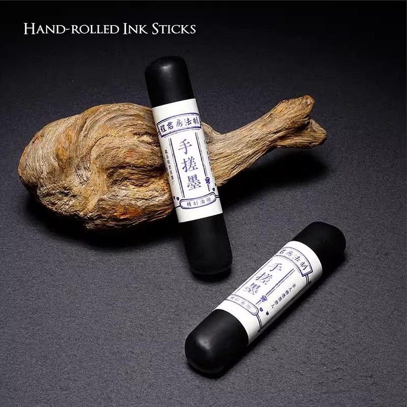 Puede incluir: Dos barras de tinta negras enrolladas a mano con etiquetas blancas con caracteres chinos. Las barras cil&iacute;ndricas descansan sobre una superficie oscura con un trozo de madera flotante. El texto "Hand-Rolled Ink Sticks" est&aacute; en la parte superior.