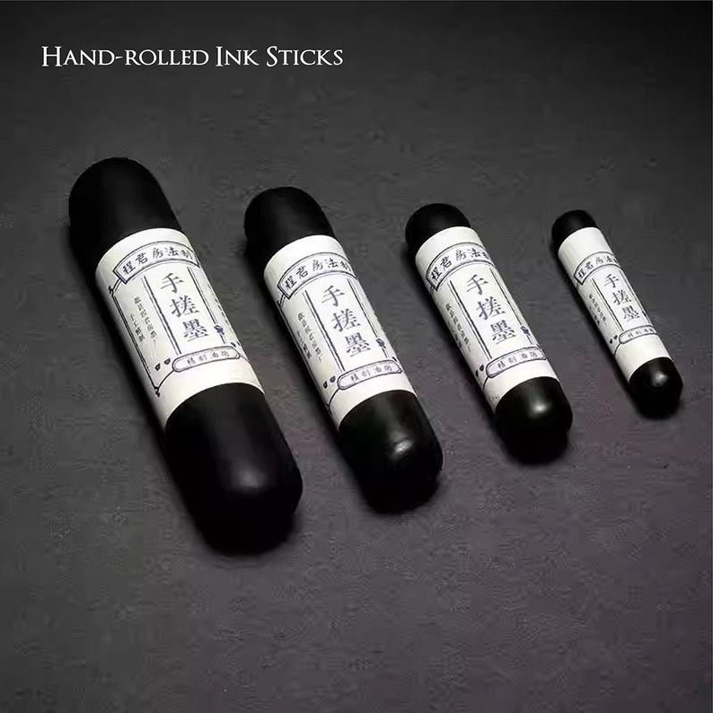 Puede incluir: Se muestran cuatro barras de tinta enrolladas a mano de diferentes tama&ntilde;os sobre un fondo oscuro. Cada barra es negra con una etiqueta blanca con caracteres chinos. El texto "HAND-ROLLED INK STICKS" est&aacute; en la parte superior de la imagen.