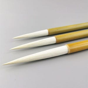 Può includere: Tre pennelli da calligrafia con manici in bambù e setole bianche. I pennelli sono disposti diagonalmente, mostrando le loro punte affusolate. I manici sono di colore marrone chiaro e le setole sono di un bianco brillante.