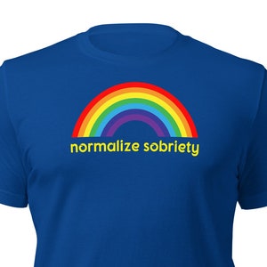 Può includere: Una t-shirt blu reale con un arcobaleno e il testo "normalizzare la sobrietà" in giallo.