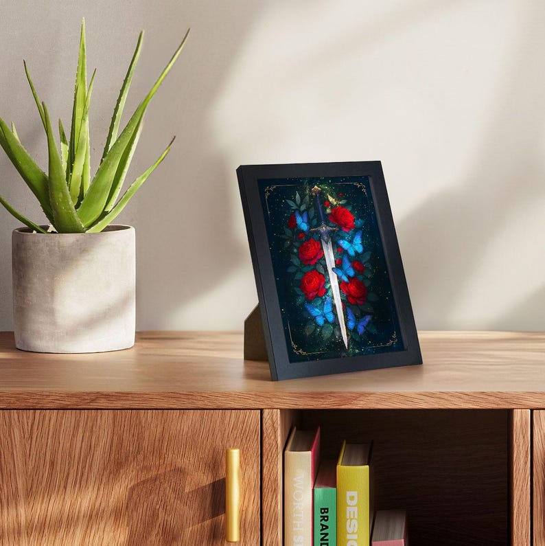 May include: A framed art print featuring a sword, red roses, and blue butterflies against a dark background. The black frame is displayed on a wooden surface, with a potted plant and books in the background. The artwork has a fantasy theme.