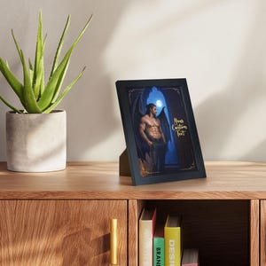 May include: A black framed print featuring a muscular figure with dark wings, set against a blue and gold background with a full moon. The text "Your Custom Text" is displayed in gold. The frame is displayed on a wooden surface.