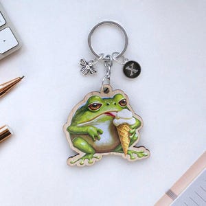 May include: A green frog keyring with a wooden base, holding an ice cream cone. The keyring has a silver chain with a bee and a silver charm with an X on it.