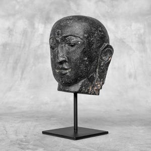Könnte beinhalten: Schwarze Steinskulptur eines Buddha-Kopfes auf einem schwarzen Metallständer. Die Skulptur ist detailliert und zeigt den ruhigen Ausdruck des Buddha.