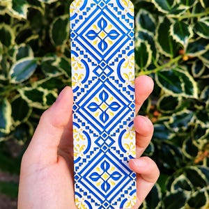 Peut inclure: Un marque-page rectangulaire arborant un motif géométrique répétitif en bleu vif, jaune éclatant et blanc. Le design se compose de losanges interconnectés et de motifs floraux complexes, offrant un aspect superposé et texturé.