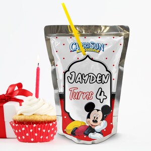 May include: A Capri Sun juice pouch with a yellow straw, featuring a Mickey Mouse design and the text "JAYDEN Turns 4". A cupcake with white frosting and a red candle is next to the juice pouch. A wrapped gift box with a red bow is also in the image.