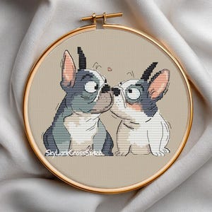 May include: A cross-stitch embroidery hoop featuring two cartoon French Bulldogs, one blue and gray, the other white and gray, nose to nose. The background is a light tan color, and the hoop is gold. The text "SkyLarkCrossStitch" is visible.