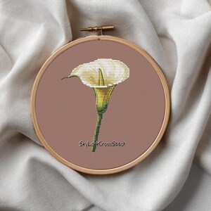 May include: A cross-stitch embroidery of a white calla lily in a wooden hoop. The flower has a yellow center and green stem, set against a muted pink background. The text "SkyLarkCrossStitch" is at the bottom.