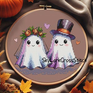 Puede incluir: Bordado de punto de cruz de dos figuras de fantasmas en un aro de madera. Un fantasma lleva una corona de calabaza y un velo, el otro un sombrero de copa. El diseño incluye corazones y el texto "SkyLarkCrossStitch".
