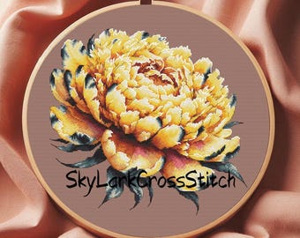 May include: A cross-stitch embroidery of a yellow peony flower with dark blue and black accents, set in a wooden hoop. The text "SkyLarkCrossStitch" is stitched below the flower on a brown background.