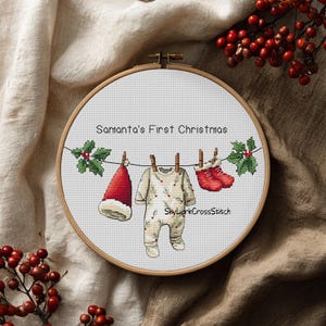 May include: A circular wooden embroidery hoop containing a cross-stitch pattern of a clothesline with a Santa hat, a baby onesie with a pine needle print, and red baby booties. The text reads Samanta's First Christmas and SkyLarkCrossStitch.