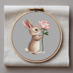 Pode incluir: Bordado em ponto cruz de um coelho segurando uma flor rosa. O coelho é marrom claro com o interior das orelhas rosa, em um fundo cinza. O bordado é emoldurado em um bastidor de madeira, com o texto "SkyLarkCrossStitch" na parte inferior.