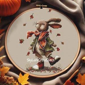 Può includere: Ricamo a punto croce di un coniglio con sciarpa e cappotto, che cammina tra le foglie che cadono. Il coniglio è in un telaio da ricamo in legno. Il testo "SkyLarkCrossStitch" è nella parte inferiore dell'immagine. La tavolozza dei colori include sfumature di marrone, verde e arancione.