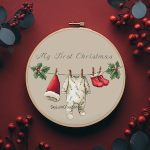 May include: A round wooden embroidery hoop containing a cross-stitch pattern on beige fabric. The design features a clothesline with a red hat, a white onesie with green sprigs, and red booties. Text reads My First Christmas and SkyLarkCrossStitch.