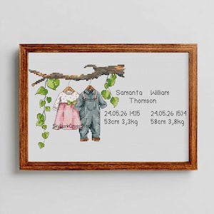 May include: A framed cross-stitch artwork featuring two baby outfits hanging from a branch with green leaves. The pink dress and blue overalls are accompanied by the names Samanta and William Thomson, along with their birth details: 24.05.26, 53cm, 3,3kg and 58cm, 3,8kg.