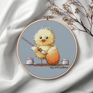 May include: Cross-stitch embroidery in a wooden hoop features a yellow chick painting an orange egg. Two small paint buckets are on either side. The text "SkyLarkCrossStitch" is at the bottom.