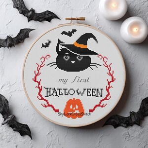 Peut inclure: Une broderie au point de croix dans un cercle en bois représentant un chat noir avec un chapeau de sorcière à bande orange, des chauves-souris, une couronne de branches rouges, une citrouille orange et le texte 'my first HALLOWEEN'.