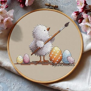 May include: Cross-stitch embroidery in a wooden hoop features a white chick holding a paintbrush, poised to decorate Easter eggs. The eggs are yellow, pink, orange, and blue with various patterns. The text "SkyLarkCrossStitch" is visible.