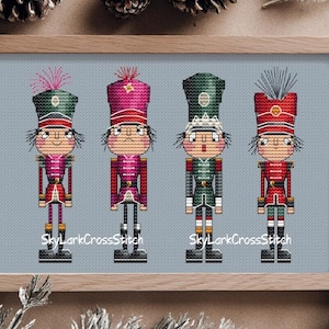 Op de afbeelding: Kruissteekkunstwerk met vier speelgoedsoldaat-notenkrakers in een houten frame. Elke notenkraker heeft een verschillend gekleurd uniform, waaronder rood, roze en groen. De tekst "SkyLarkCrossStitch" is zichtbaar onderaan het kunstwerk.