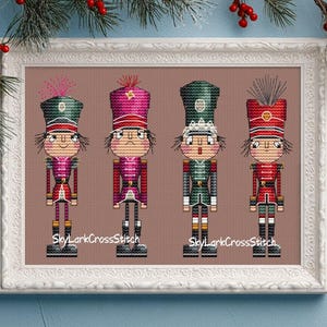 Puede incluir: Obra de arte de punto de cruz con cuatro cascanueces soldados de juguete en un marco blanco. Cada cascanueces tiene un uniforme de diferente color, incluyendo rojo, verde y rosa. La obra de arte está decorada con vegetación navideña.