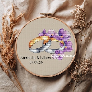 Peut inclure: Un cercle de broderie au point de croix avec deux anneaux entrelacés, l'un doré et l'autre argenté, ornés de fleurs violettes. Les noms "Samanta & William" et la date "24.05.26" sont brodés en dessous. Le fond est beige neutre.