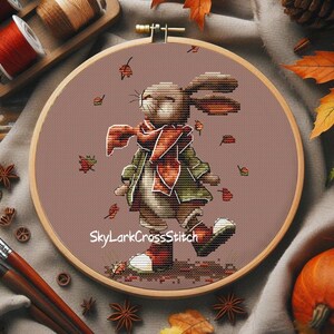 May include: A cross-stitch embroidery in a wooden hoop depicts a rabbit in a scarf and coat, walking amongst falling leaves. The design uses brown, green, and orange hues. The text "SkyLarkCrossStitch" is visible.