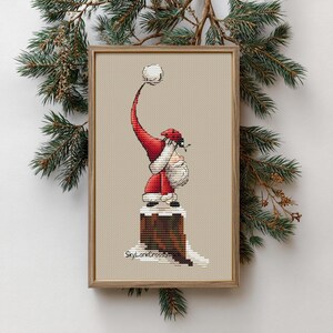 May include: Cross-stitch art of Santa Claus in a red suit and hat, holding a white ball. The artwork is framed in a wooden frame and set against a neutral background. The image is decorated with pine branches.