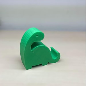 Könnte beinhalten: Ein grüner, dinosaurierförmiger Handyständer. Der Ständer ist aus Kunststoff und hat eine strukturierte Oberfläche.