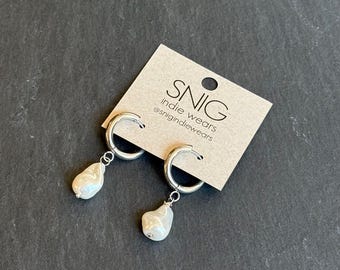 Könnte beinhalten: Silberne Creolen mit einem baumelnden Perlen-Anhänger. Die Ohrringe befinden sich auf einer Karte mit der Aufschrift "SNIG indie wears @snigindiewears".