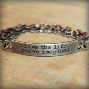 May include: A metal bracelet with a curved rectangular plate engraved with the words "live the life you've imagined." The bracelet features multiple chains in shades of silver, copper, and bronze, with gold-colored clasps.