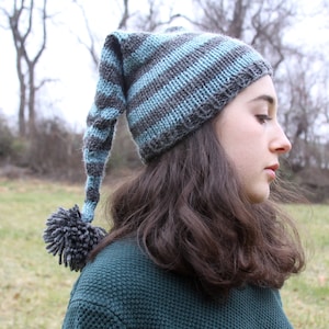 May include: A person wearing a blue and gray striped knit hat with a long, pointed top and a pom-pom on the end. The hat is pulled down over their forehead and the person is looking off to the side.