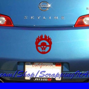 May include: A blue Nissan Skyline car with a red Mad Max Fury Road skull and flames decal on the back bumper. The car has a California license plate and a Nismo emblem.