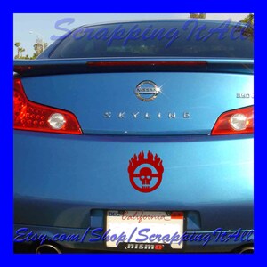 May include: A blue Nissan Skyline car with a red Mad Max Fury Road skull and flames decal on the back bumper. The car has a California license plate.