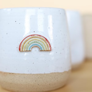 May include: White ceramic mug with a rainbow design. The mug has a speckled glaze and a beige rim.