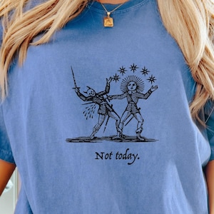 May include: A blue t-shirt featuring a black graphic of a sun and a devil in combat. The text "Not today." is printed beneath the graphic.