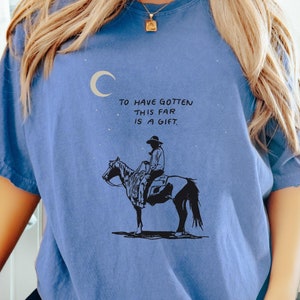 May include: A blue t-shirt with a graphic of a cowboy riding a horse under a crescent moon. The text on the shirt reads "To have gotten this far is a gift."