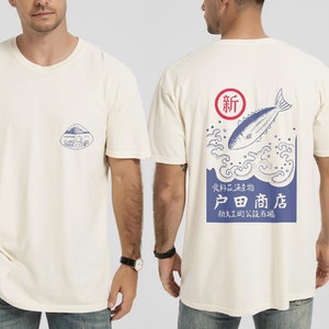 以下が含まれることがあります： 白地に青と白のグラフィックがプリントされたTシャツ。グラフィックには魚、波、そして「新 食料品海産物 户田商店 新大工町公設市場」の文字が描かれている。
