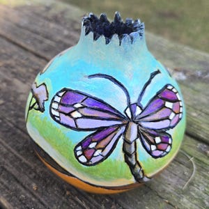 May include: A hand-painted, gourd-shaped vase with a dragonfly design. The vase features a light blue background with a large purple and white dragonfly. The bottom is orange, and the top has a jagged edge.