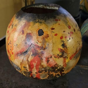 May include: A round, hand-painted gourd with a black, brown, yellow, red, and orange design. The gourd has a large opening at the top and is decorated with abstract shapes and figures.