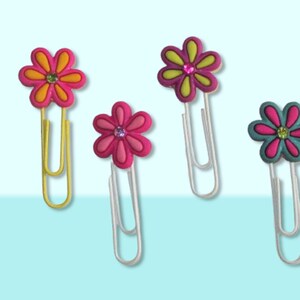 Four colorful flower-shaped paper clips. The paper clips are yellow, pink, green, and blue. Each paper clip has a flower design with a rhinestone in the center.