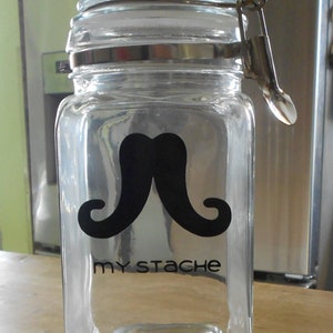 May include: A clear glass jar featuring a black mustache design and the words "my stache". The jar has a hinged lid with a metal clasp. The rectangular jar is on a wooden surface.